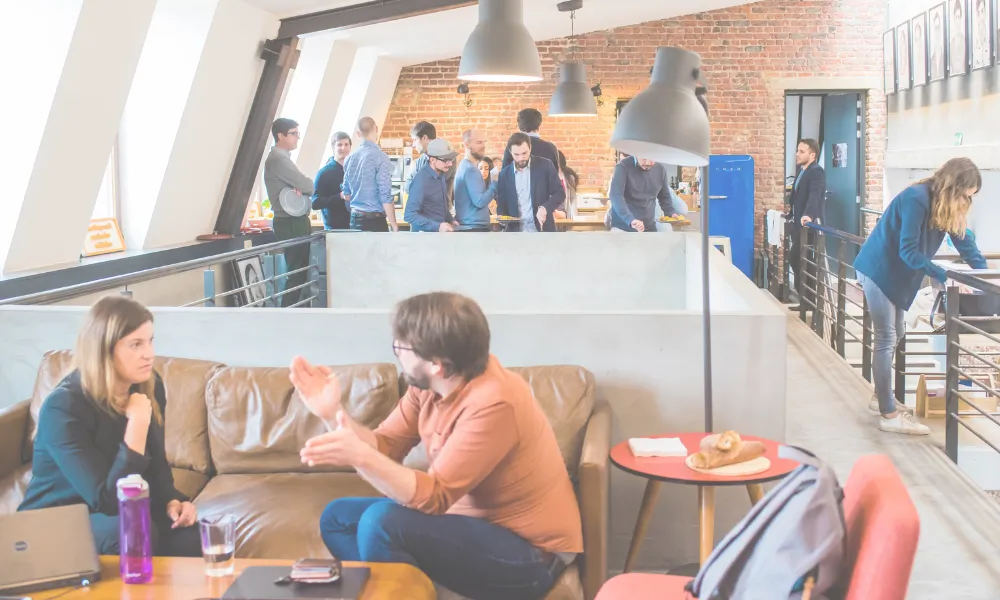 An office setting where two people are talking and people in the background.